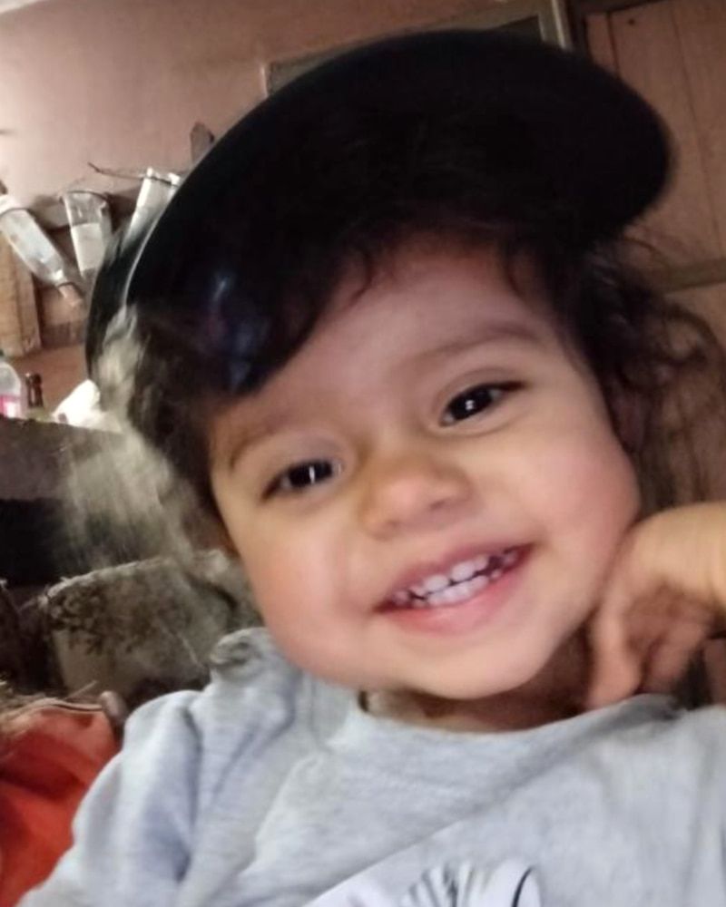 Mia, de 2 años, sonríe radiante con una gorra negra y su manito apoyada en la mejilla.